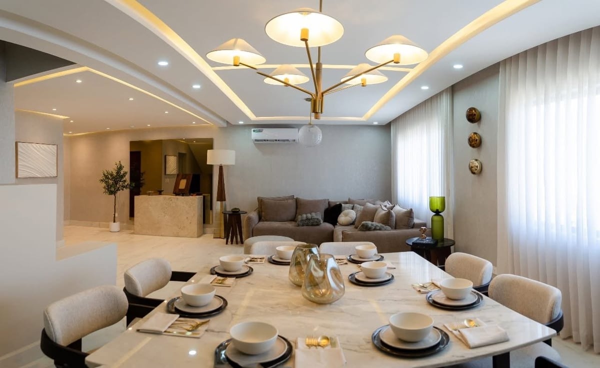 Modern dining room with round table set for six, cream upholstered chairs, geometric ceiling lights, and living area visible in background