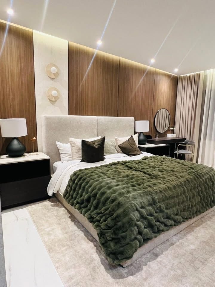 Modern luxury bedroom with green quilted bedspread, beige upholstered headboard, black nightstands, wood wall paneling, and recessed ceiling lights