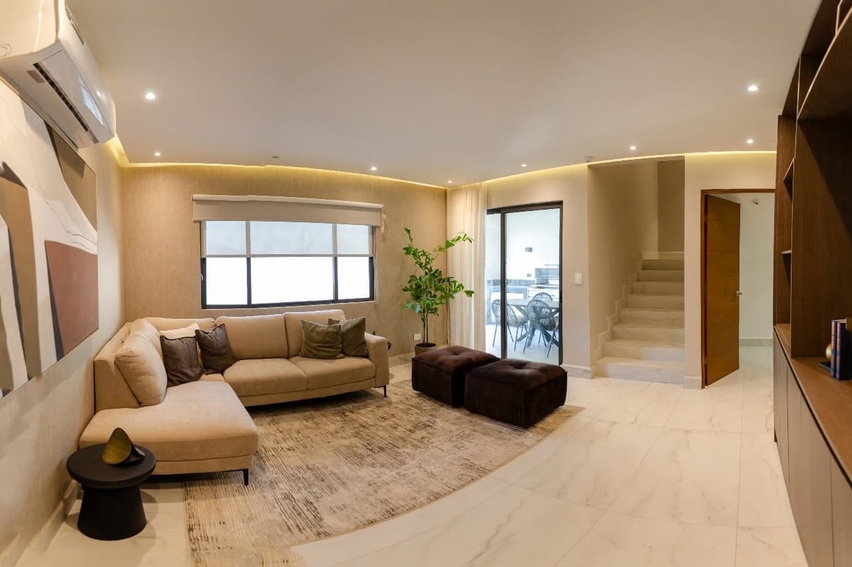 Modern living room with beige sofa, dark ottoman, warm ceiling lights, large windows, and neutral decor throughout