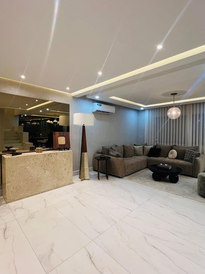 Modern apartment interior with beige reception desk, marble flooring, gray sectional sofa, and contemporary ceiling lighting